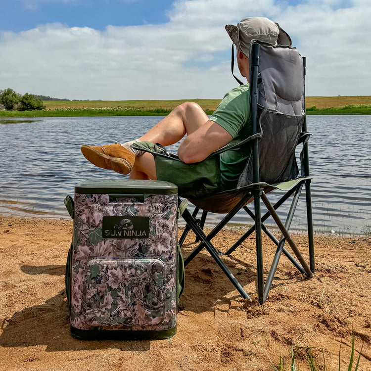 Person sitting in a camping chair by a lake with a camouflage cooler bag next to them.