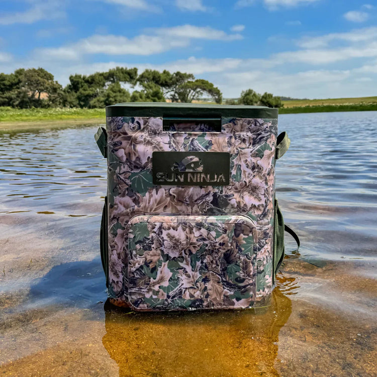 Camouflage-patterned cooler bag with 'Fishing Ninja' branding on a lakeshore.
