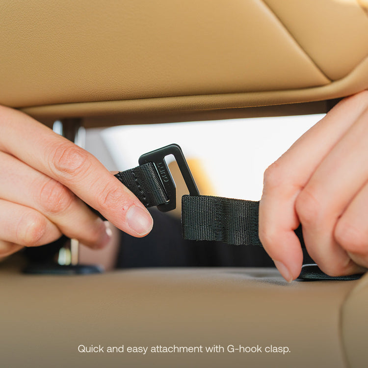 Close-up of hands fastening a black strap with a G-hook clasp on a tan car seat, showcasing the Luno® Seatback Organizer. The image text reads: "Quick and easy attachment with G-hook clasp for seamless vehicle organization or essential car camping setup.