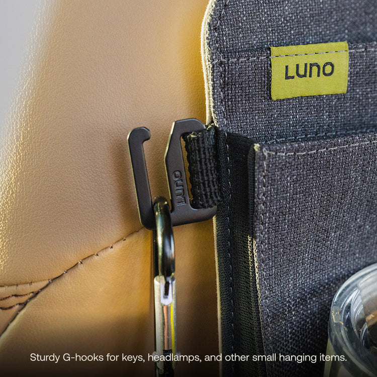 Close-up of a G-hook on the Luno® Seatback Organizer clipped to a beige car seat, displaying its "Luno" label. A keychain is attached, with a zippered pocket and plastic bottle partially visible, ideal for organizing keys, headlamps, and small items in your vehicle.