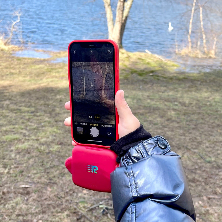 Person holding a red phone case with a phone displaying a camera interface, outdoors by a body of water.