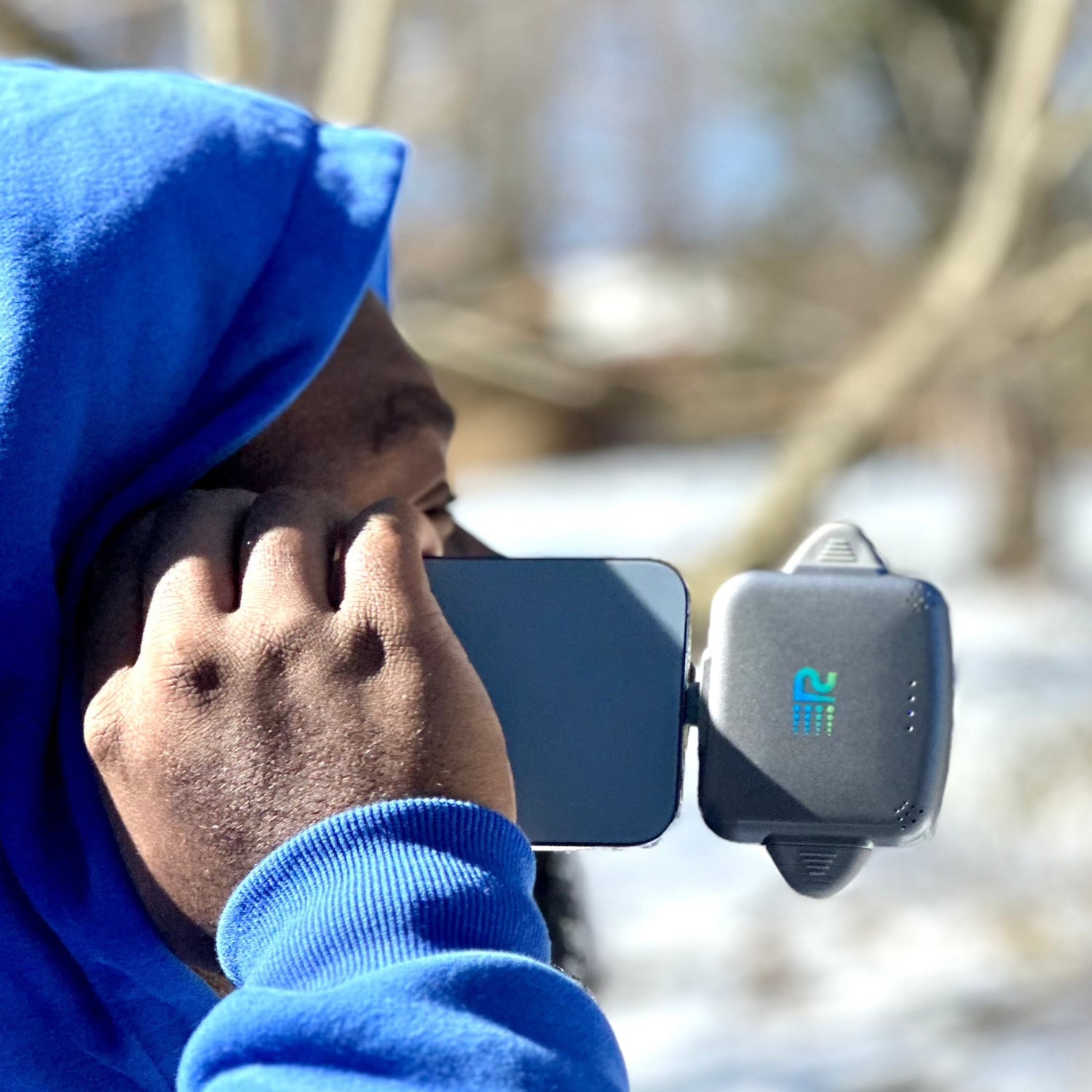 Person wearing a blue hoodie using a black and gray device outdoors.