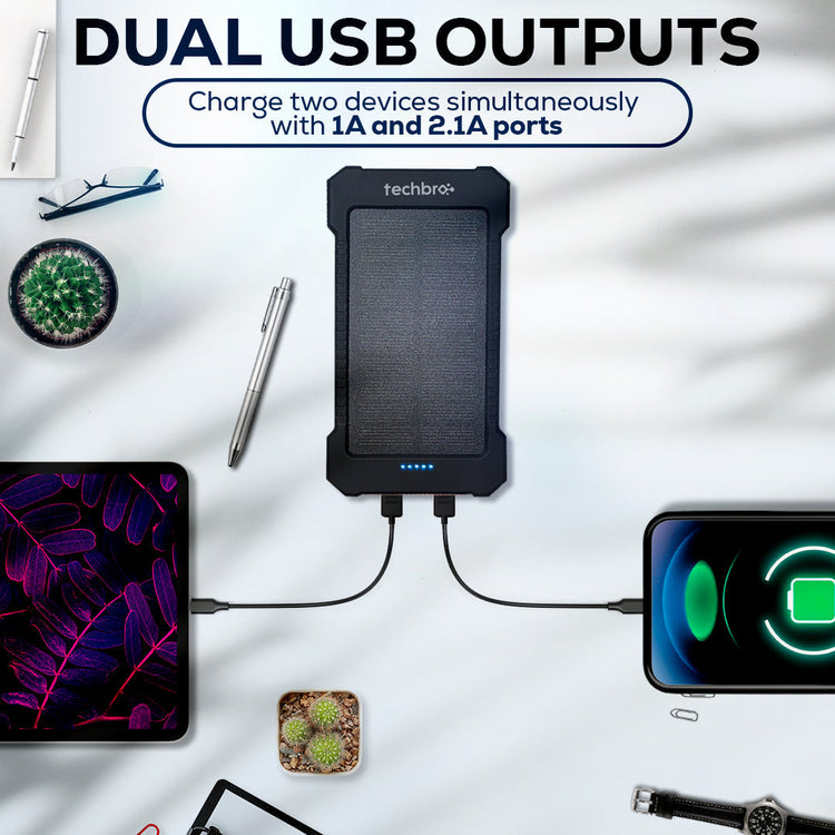 Power bank with dual USB outputs charging a tablet and phone on a white surface with office items.