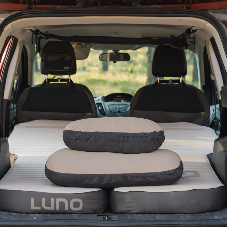 Two Luno® Packable Camping Pillows rest on an air mattress in the back of a car with the rear seats folded down, while an outdoor scene is visible through the front windshield.
