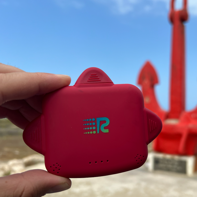 Red device with a logo held by a hand against a blue sky and playground background