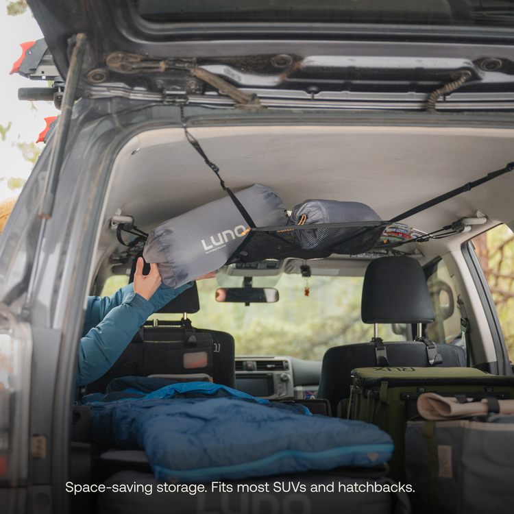 A person is storing a Luno® Cargo Hammock, in gray, hanging from the ceiling of an SUV's trunk. The trunk is filled with camping gear, including a blue sleeping bag and green bags. The text reads, "Space-saving gear storage for your adventure mobile. Fits most SUVs and hatchbacks.