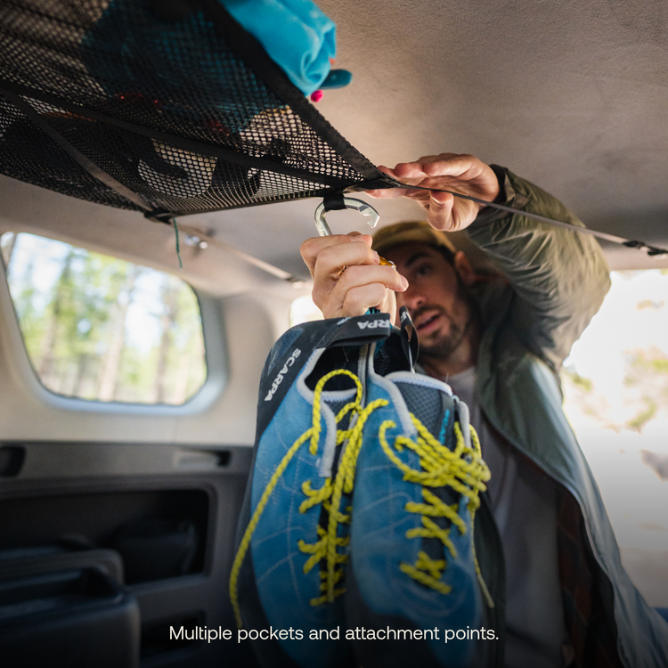 A person reaches up to access the Luno® Cargo Hammock inside their adventure mobile, attaching a pair of blue climbing shoes with yellow laces to an overhead hook. The forested scenery outside adds to the vibe. The image caption reads "Multiple pockets and attachment points for ultimate gear storage.