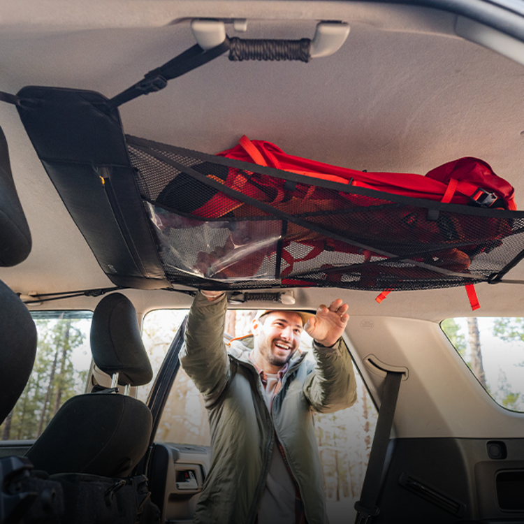 A man positions a red backpack on a Luno® Cargo Hammock, which is attached to the ceiling of his adventure mobile. The surrounding area, visible through the car windows, is a lush forest. Smiling and dressed in a gray jacket and white hat, he appears ready for an exciting journey.