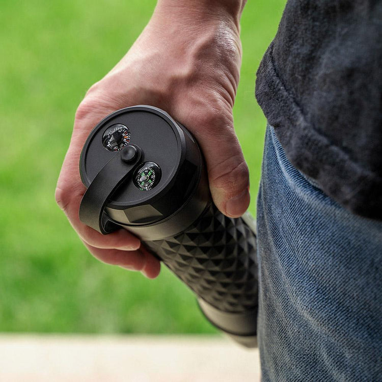 Hand holding a black and gray device with a textured grip against a green outdoor background
