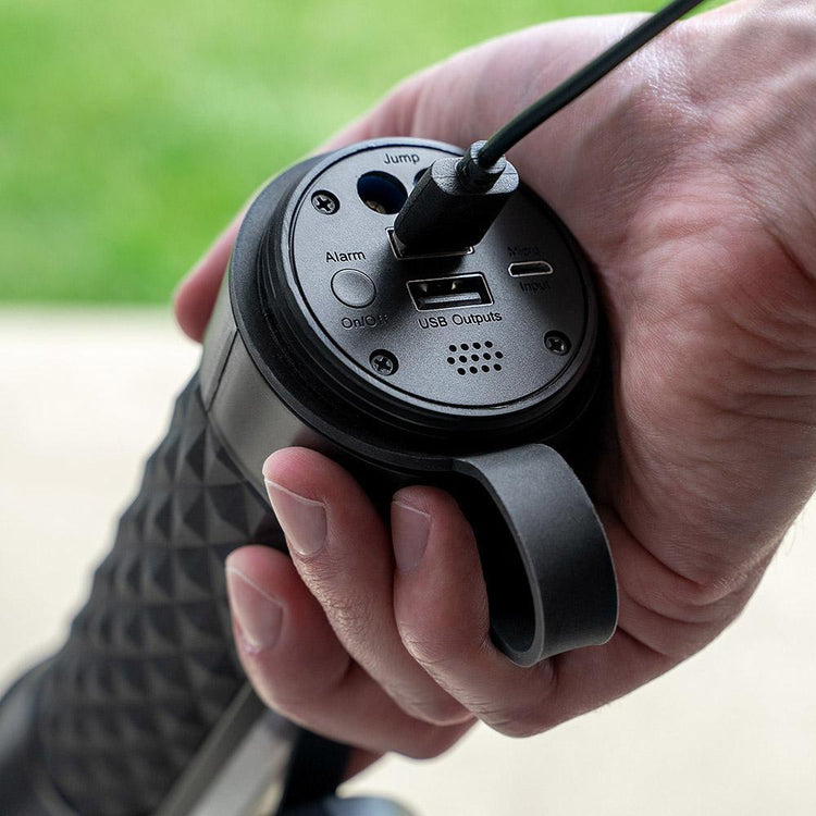 Hand holding a black electronic device with USB ports and buttons, outdoors.