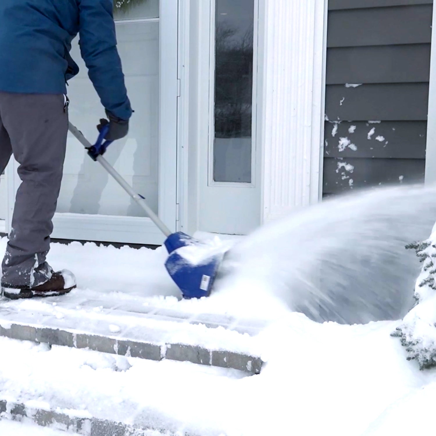 Person using a snow blower to clear snow from a driveway in front of a house.