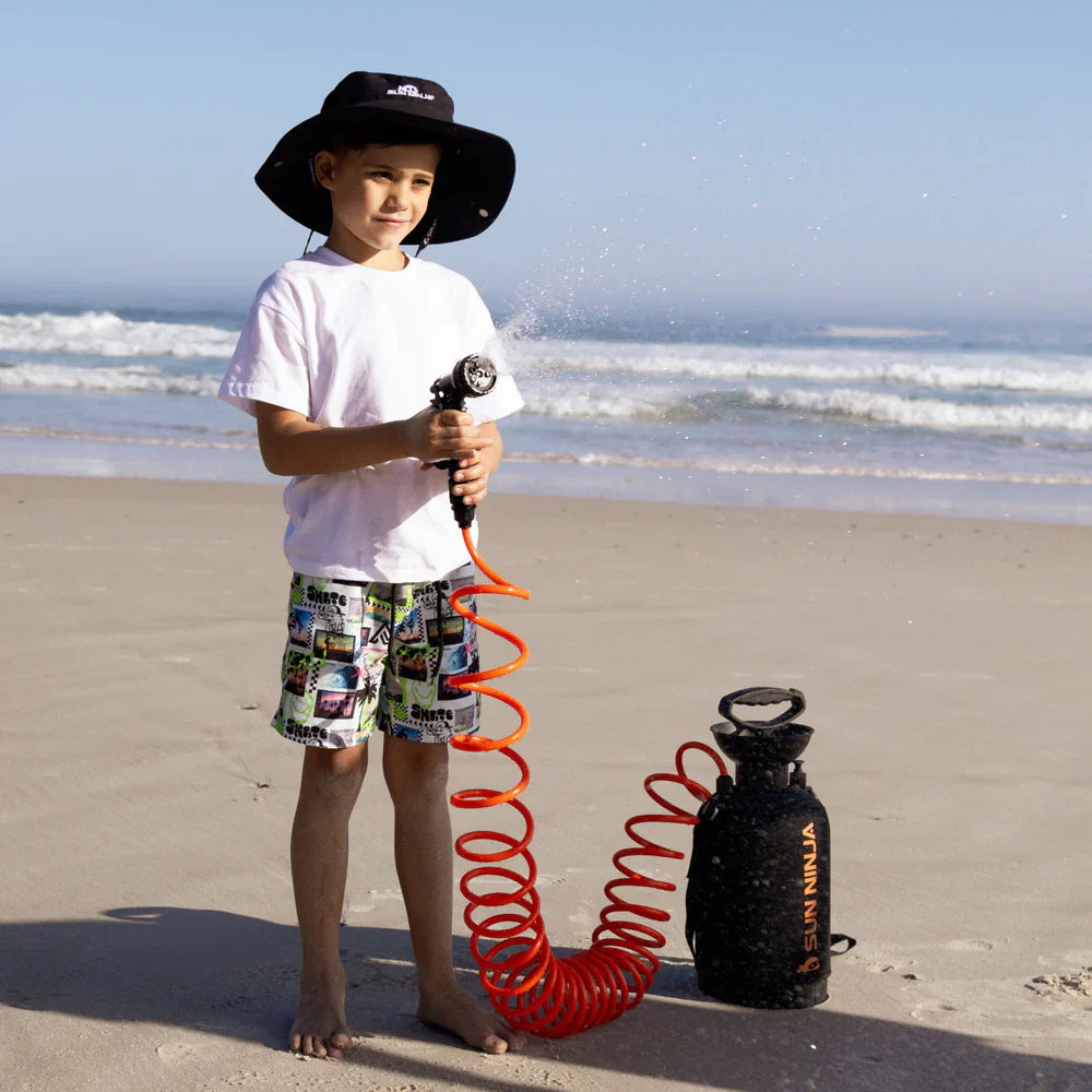 Child on a beach holding a water gun with a coiled hose, wearing a black hat and colorful shorts.