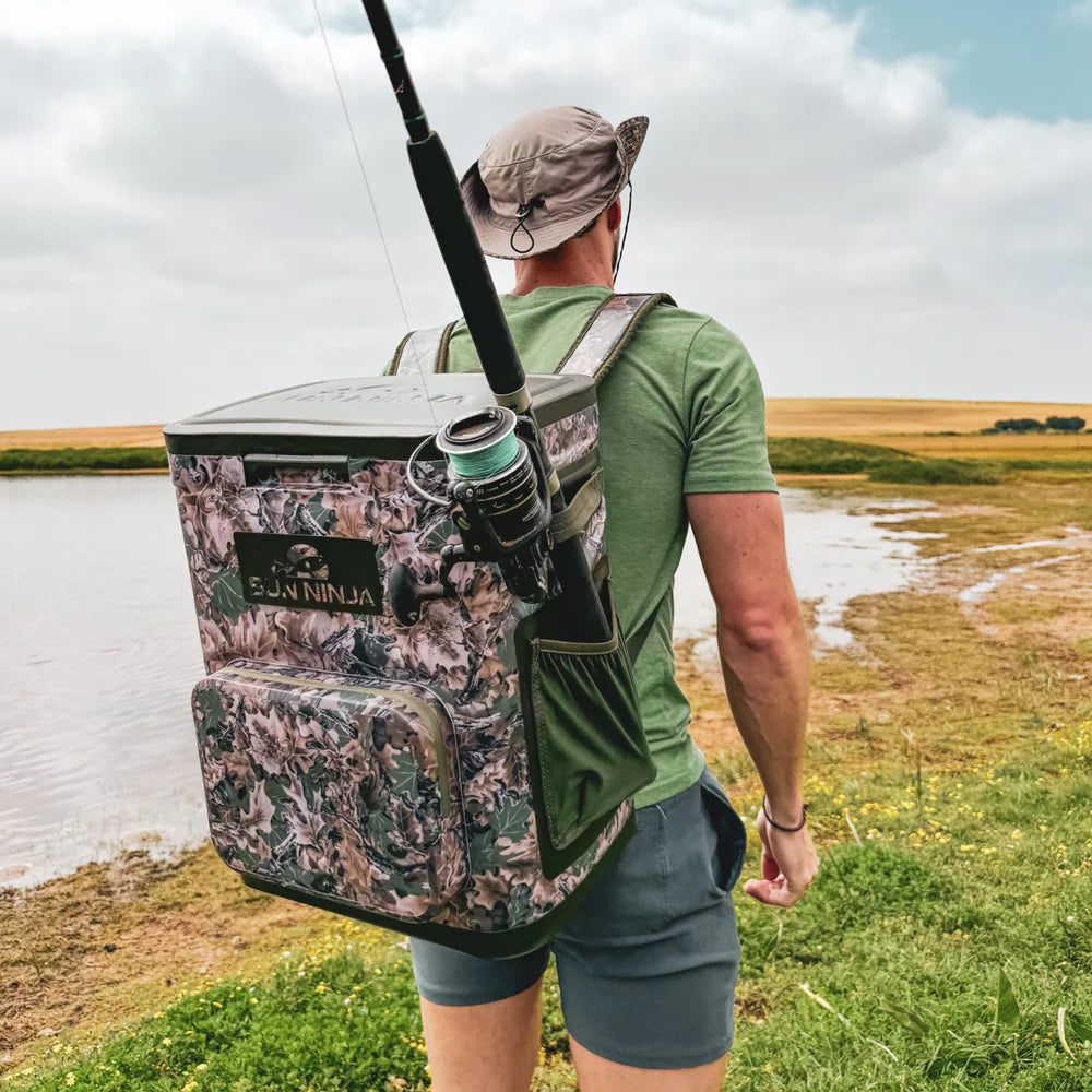 Person with a camouflage backpack holding a fishing rod, standing by a body of water.