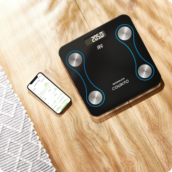 Digital body fat scale on a wooden surface with a smartphone displaying a fitness app.