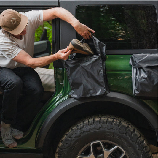 A person puts dirty running shoes with red laces into a black Luno® Gear & Shoe Storage Bag that's attached to the side of a car.