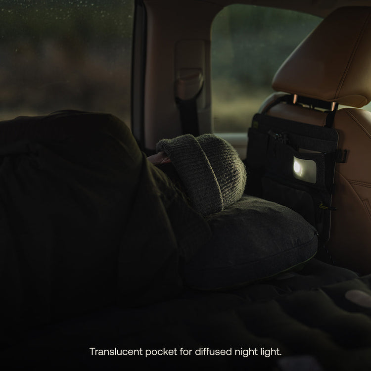 A beanie-wearing person sleeps on a pillow in a car's backseat, ideal for camping. A dim light glows from Luno® Seatback Organizer's translucent pocket attached to the front seat, enhancing the cozy ambiance. Text reads, "Translucent pocket for diffused night light.