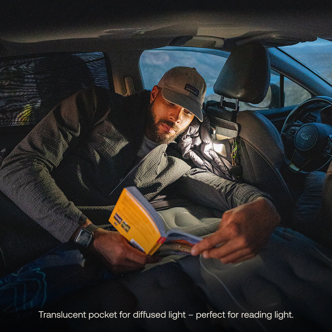 Reclining in the backseat with a cap and dark jacket, one reads by a translucent light, showcasing the cozy interior with a Luno® Seatback Organizer for efficient organization and a sleeping bag: "Translucent pocket provides diffused light—ideal for reading.