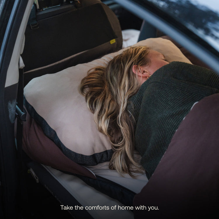 A person with blonde hair relaxes on pillows and blankets in a car’s backseat. The caption reads, “Bring home-like comfort anywhere with the Luno® Packable Camping Pillow—even on the road.”.