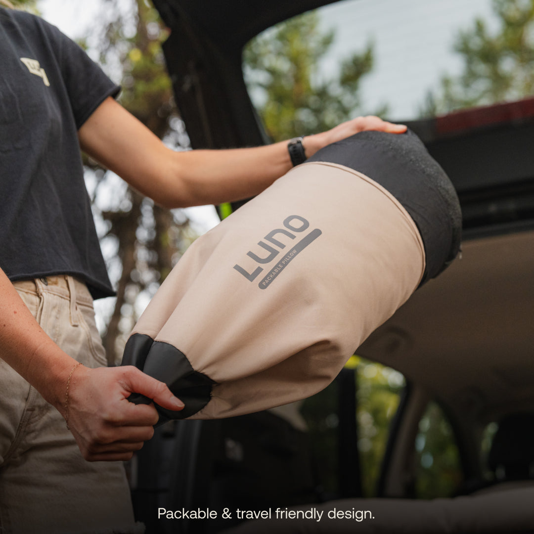 A person holds a rolled-up beige Luno® Packable Camping Pillow by an open car trunk, ready for packing. Text reads, "Packable & travel friendly design." Ideal as a compact camp pillow from Luno® for your next adventure.