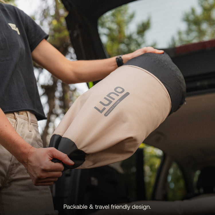 A person holds a rolled-up beige Luno® Packable Camping Pillow by an open car trunk, ready for packing. Text reads, "Packable & travel friendly design." Ideal as a compact camp pillow from Luno® for your next adventure.