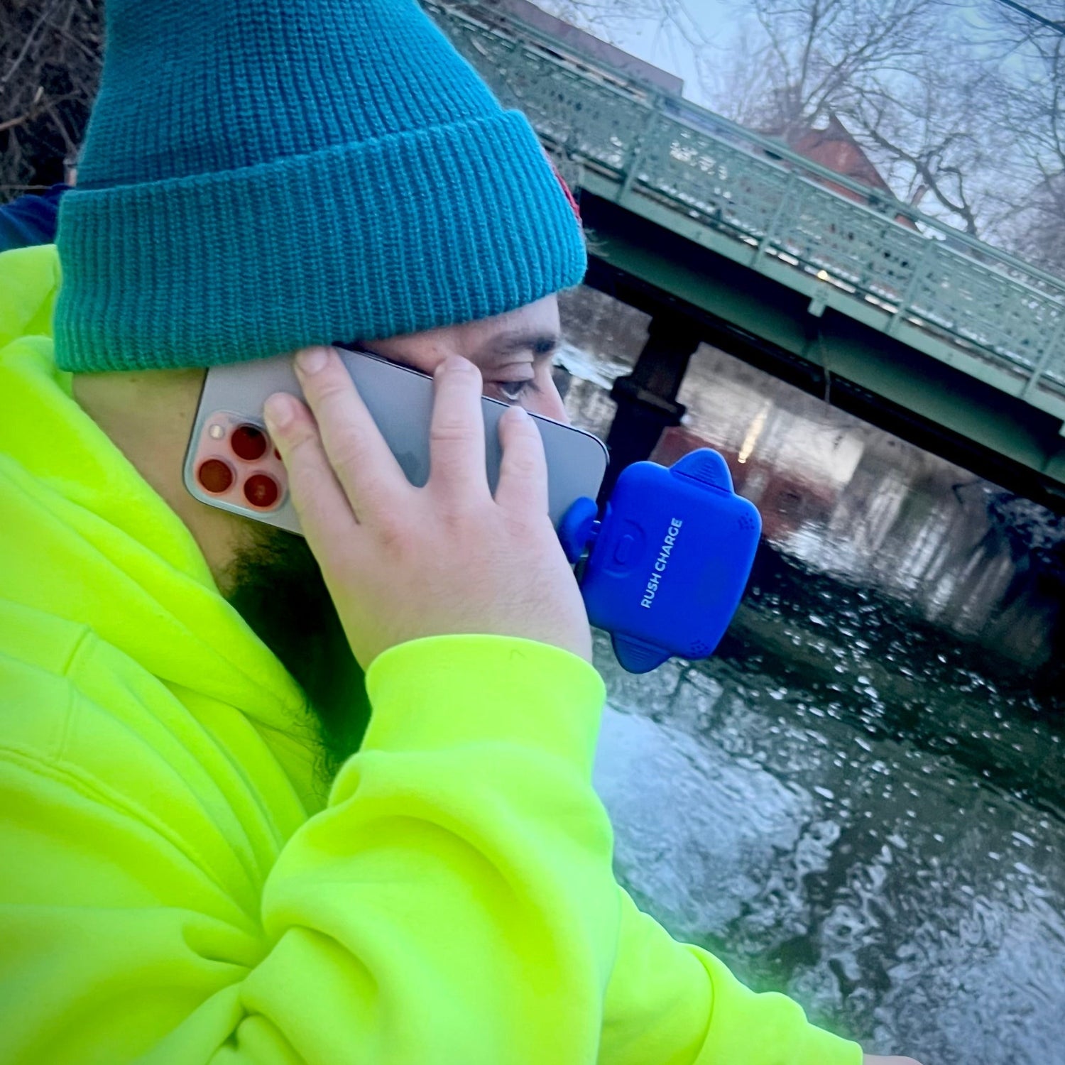 Person in bright yellow jacket and blue beanie using a phone with a blue case, standing by a body of water.