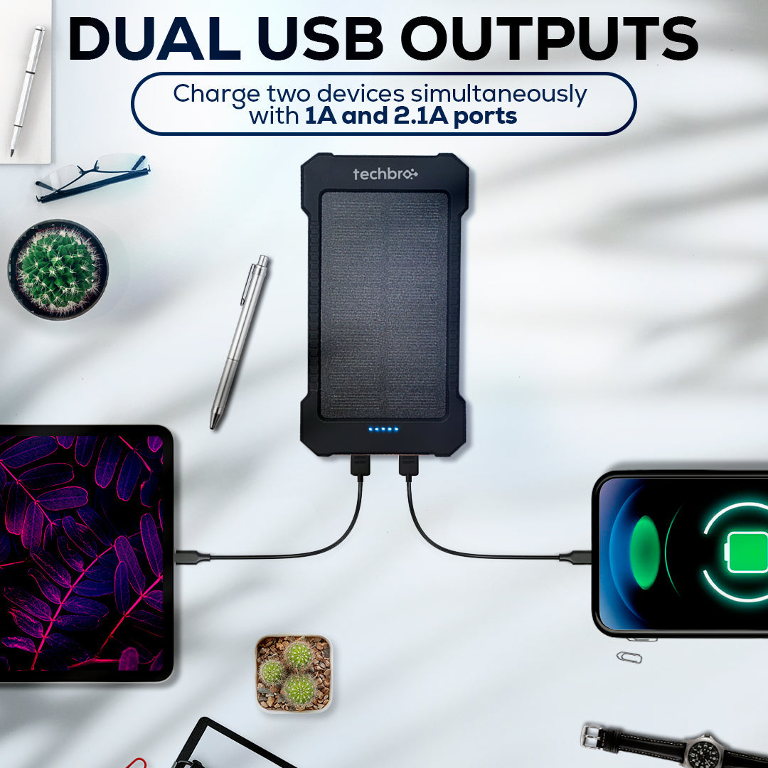 Power bank with dual USB outputs charging a tablet and phone on a white surface with office items.