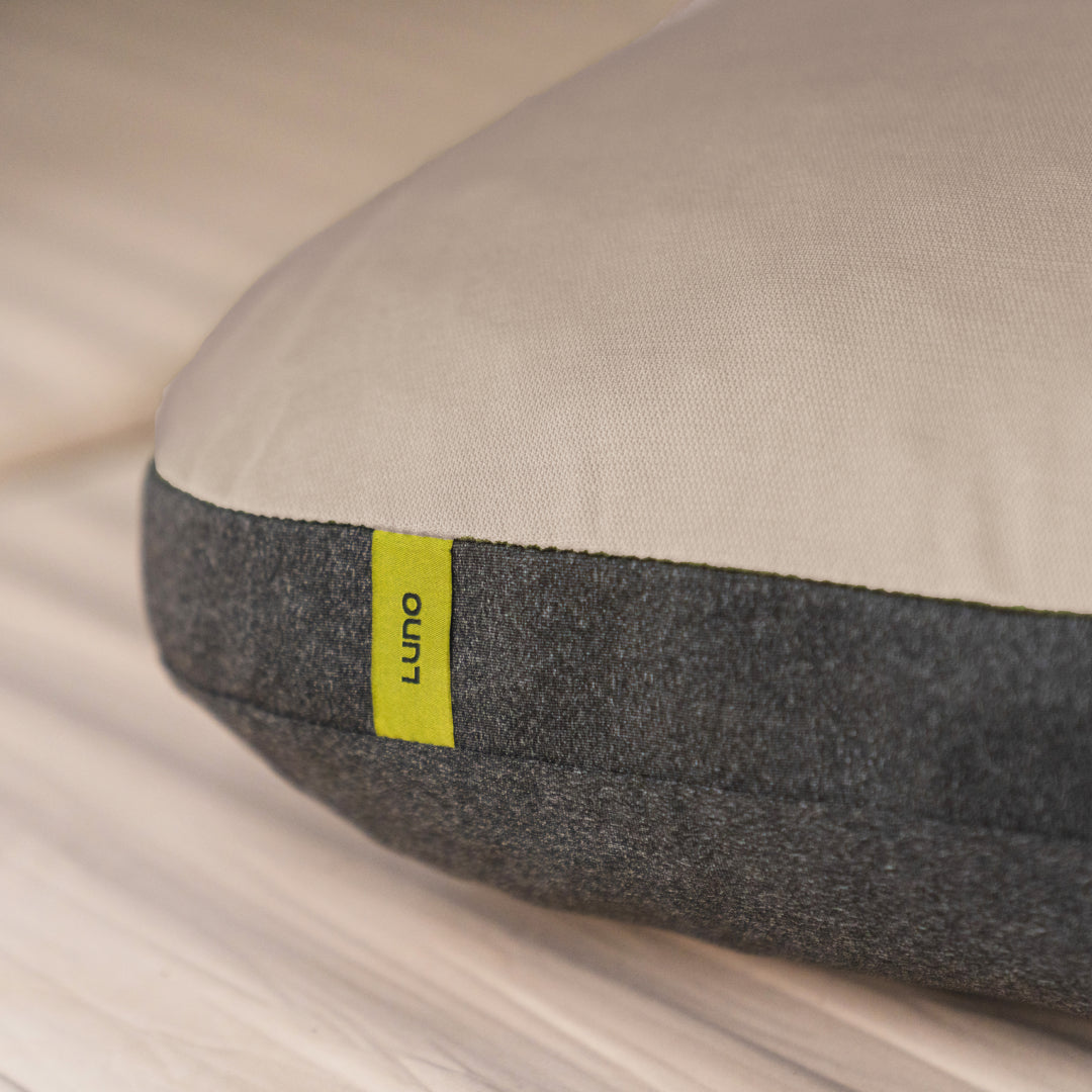 Close-up of the Luno® Packable Camping Pillow, featuring a beige top, dark gray bottom, and a small yellow Luno® tag, displayed on a wooden surface.