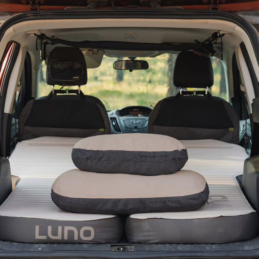 A car’s back seat is converted into a sleeping area with a gray inflatable mattress and Luno® Packable Camping Pillow. The mattress features “DRIVE. CAMP. EXPLORE.”.