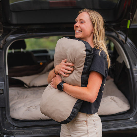 A car’s back seat is converted into a sleeping area with a gray inflatable mattress and Luno® Packable Camping Pillow. The mattress features “DRIVE. CAMP. EXPLORE.”.