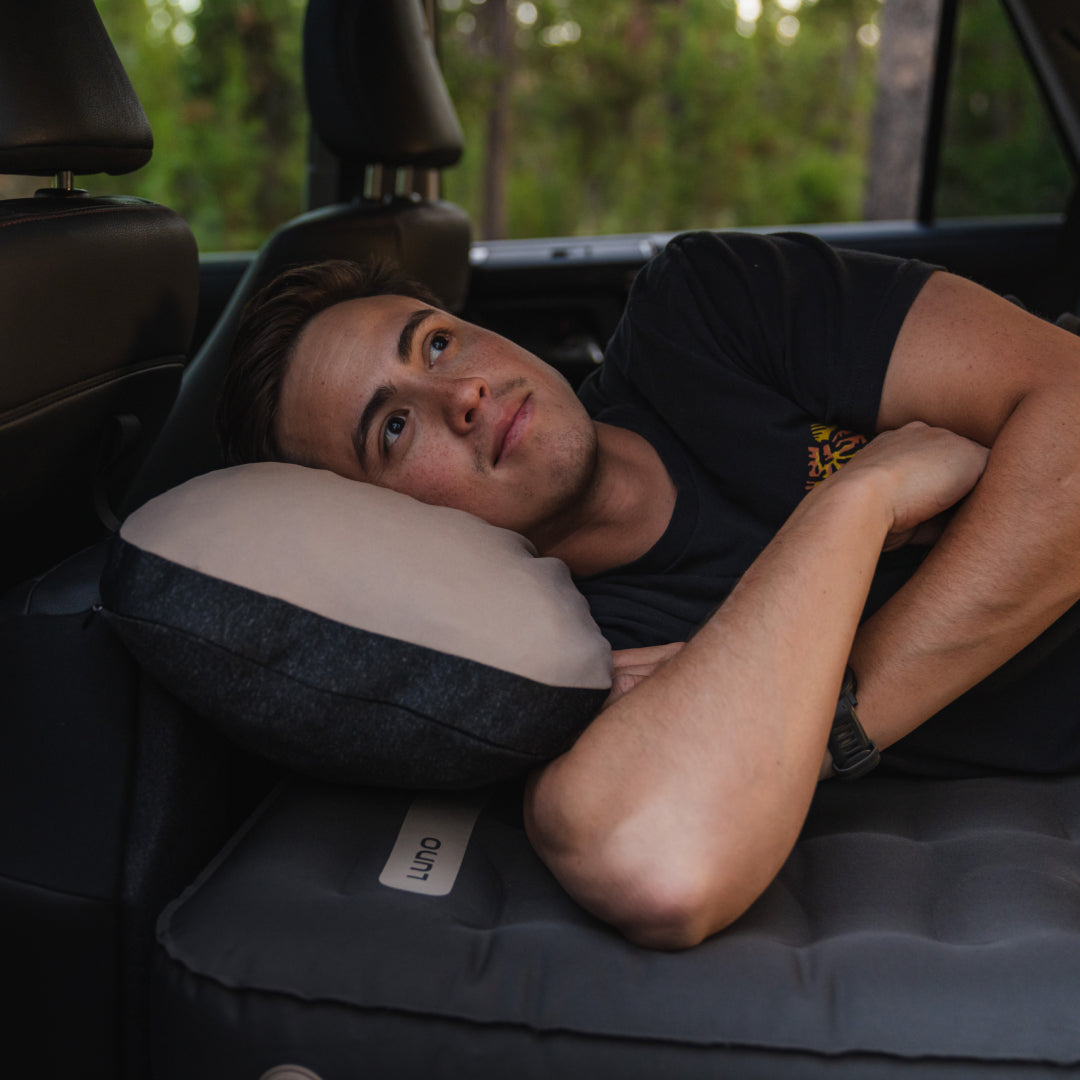A young man reclines on an inflatable mattress in the back of a car, resting his head on a Luno® Packable Camping Pillow, with car seats around him and a forested backdrop visible outside the window.