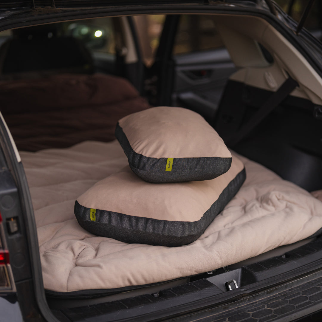 Two Luno® Packable Camping Pillows in tan and dark gray are stacked on a beige mattress in the back of an SUV with the trunk open, creating a cozy car camping or travel setup.