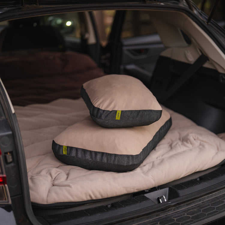 Two Luno® Packable Camping Pillows in tan and dark gray are stacked on a beige mattress in the back of an SUV with the trunk open, creating a cozy car camping or travel setup.