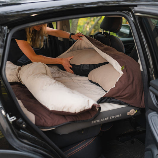 A car’s back seat is converted into a sleeping area with a gray inflatable mattress and Luno® Packable Camping Pillow. The mattress features “DRIVE. CAMP. EXPLORE.”.