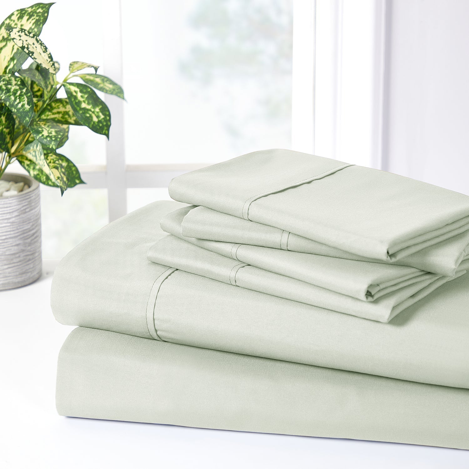 Folded light green bed sheets on a white surface with a plant in the background