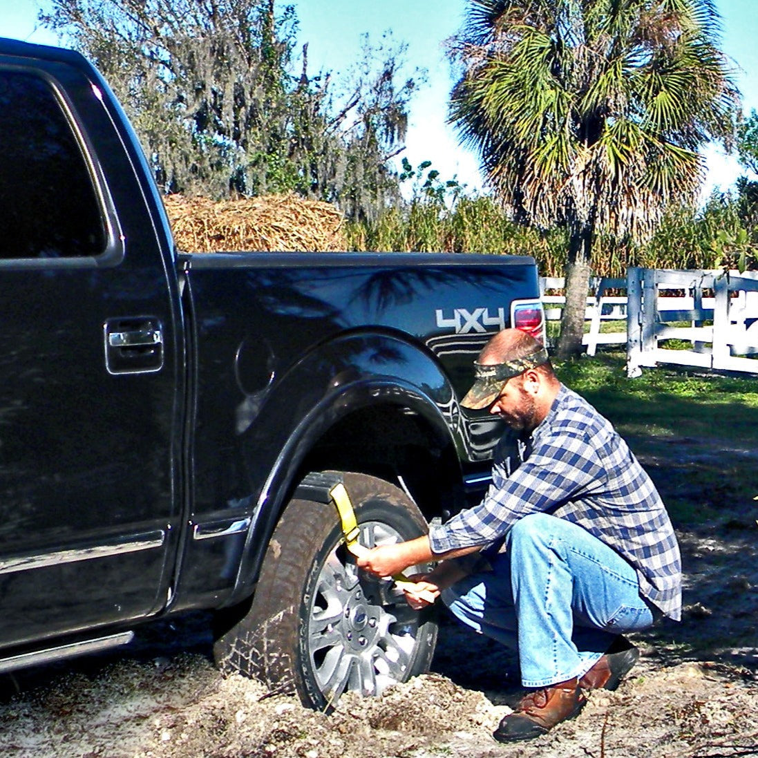 Man changing a tire on a black truck in a muddy area with trees and a white fence in the background.