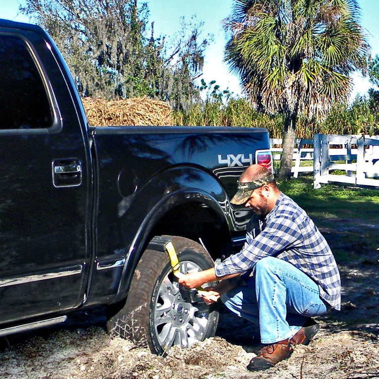 Man changing a tire on a black truck in a muddy area with trees and a white fence in the background.