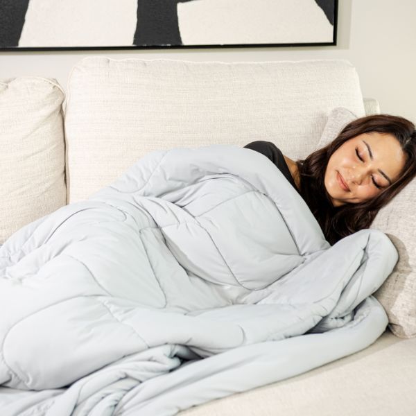 Woman lying on a couch under a light gray weighted blanket in a cozy living room.