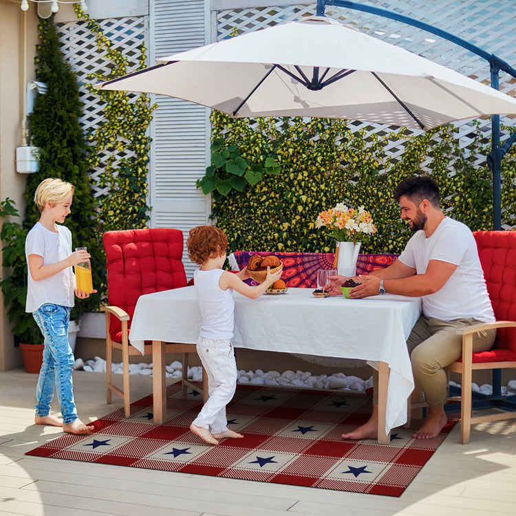 Family enjoying a meal outdoors under a large umbrella with a red and white checkered rug.