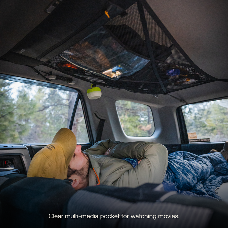 A person wearing a yellow cap and green jacket lies on their side in the backseat area of an adventure mobile, looking up at what appears to be a Luno® Cargo Hammock attached to the ceiling of the car, presumably used for holding multimedia devices for watching movies. Surrounding vegetation is visible through the windows.
