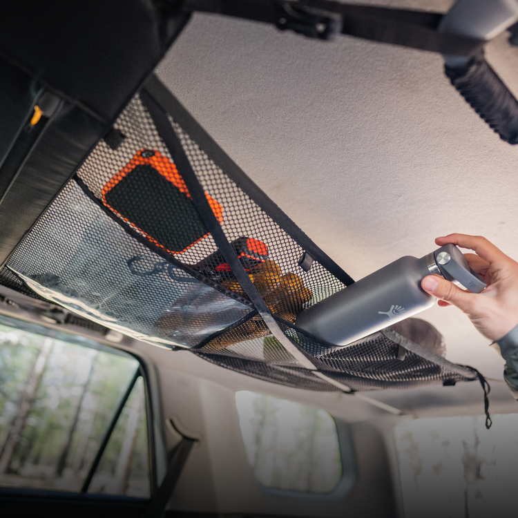 A person stores a gray water bottle in the black Cargo Hammock from Luno® inside their adventure mobile. The hammock also contains an orange item and other small objects, all securely held in this portable gear shelf. The background features a blurred view of trees visible through the vehicle's windows.