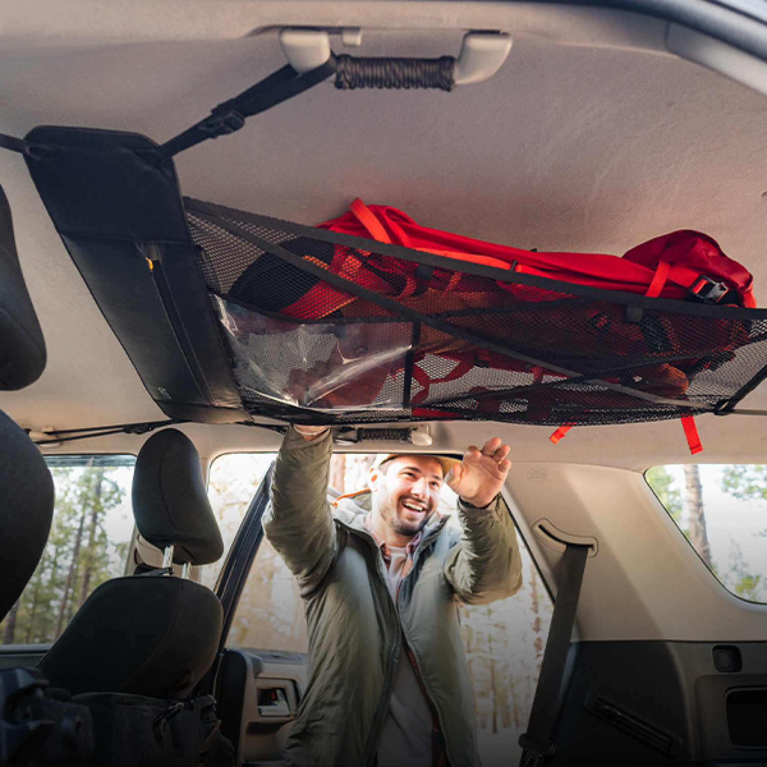 A man positions a red backpack on a Luno® Cargo Hammock, which is attached to the ceiling of his adventure mobile. The surrounding area, visible through the car windows, is a lush forest. Smiling and dressed in a gray jacket and white hat, he appears ready for an exciting journey.