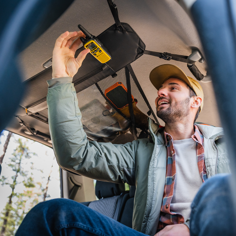 A man in a green jacket and beige cap is sitting inside an adventure mobile, reaching up to place a yellow walkie-talkie onto the Luno® Cargo Hammock, a black mesh storage net attached to the car ceiling. Trees can be seen through the car windows in the background.