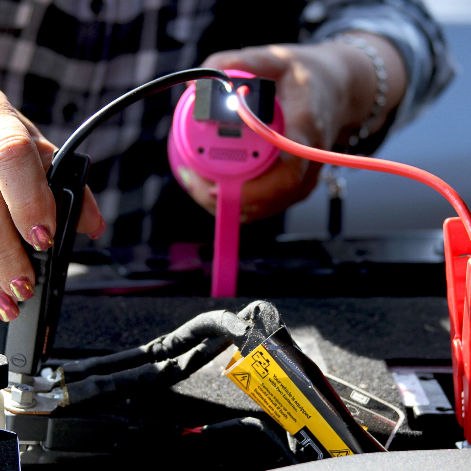 Person using a jump starter to charge a car battery with a pink phone in the background.